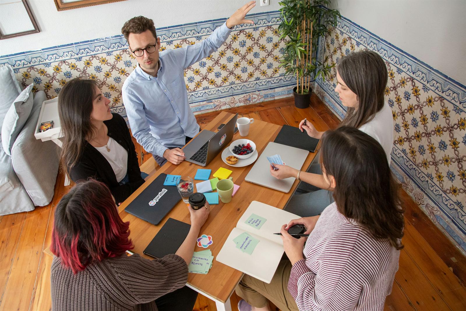 Equipe de quatro pessoas reunida em volta de uma mesa, com post-its, cadernos e laptops, enquanto um participante explica uma ideia